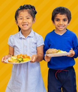 Two smiling children wearing school uniform and each holding a plate of food